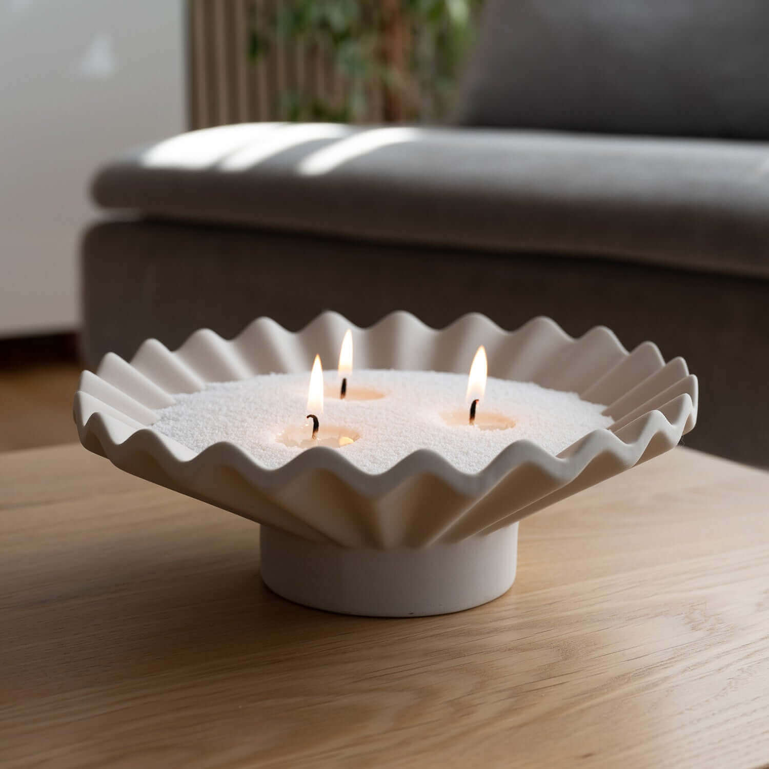 Bougie design Enora-Bougie dans un bol blanc plissé avec sable végétal blanc, trois mèches allumées, posée sur une table en bois pour une ambiance chic et naturelle.
